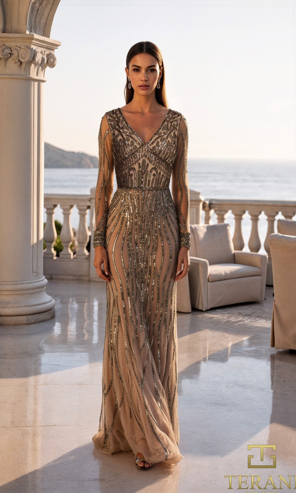 A woman in a long, sheer, beaded Champagne Silver gown (Terani 261M6727) stands on a marble terrace with columns, overlooking a coastal sunset.